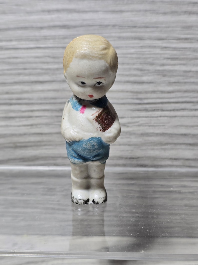 Puede incluir: Peque&ntilde;a figura de porcelana vintage de un ni&ntilde;o. La figura est&aacute; pintada con una camisa blanca, pantalones cortos azules y un libro marr&oacute;n. El ni&ntilde;o tiene el pelo rubio y una expresi&oacute;n ligeramente triste. La figura est&aacute; de pie.