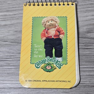 May include: A spiral-bound notepad with a yellow cover featuring a Cabbage Patch Kids graphic. The graphic shows a doll wearing a red jacket, jeans, and white shoes. Text reads "There's no one else like me..." and "Cabbage Patch Kids."
