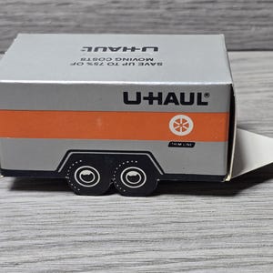 May include: A small U-Haul trailer box in silver and orange. The box displays the U-Haul logo and the text "SAVE UP TO 75% OF MOVING COSTS". The trailer has black wheels and a white trim line. The box is a miniature model.