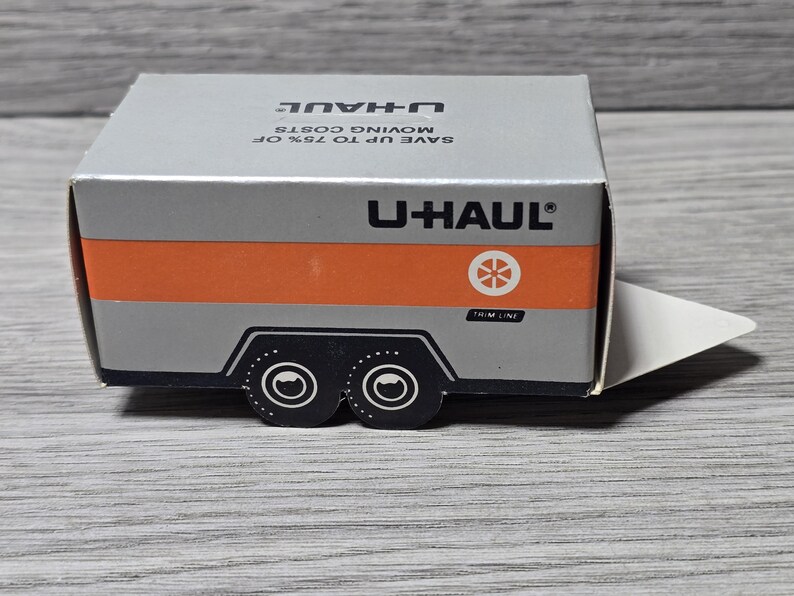 May include: A small U-Haul trailer box in silver and orange. The box displays the U-Haul logo and the text "SAVE UP TO 75% OF MOVING COSTS". The trailer has black wheels and a white trim line. The box is a miniature model.