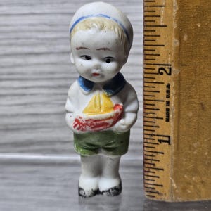 Pu&ograve; includere: Piccola statuina vintage in porcellana di un marinaio. La statuina indossa un cappello e una camicia bianchi con colletto blu, pantaloncini verdi e tiene in mano una barca rossa e gialla. La statuina &egrave; alta circa 6,35 cm.