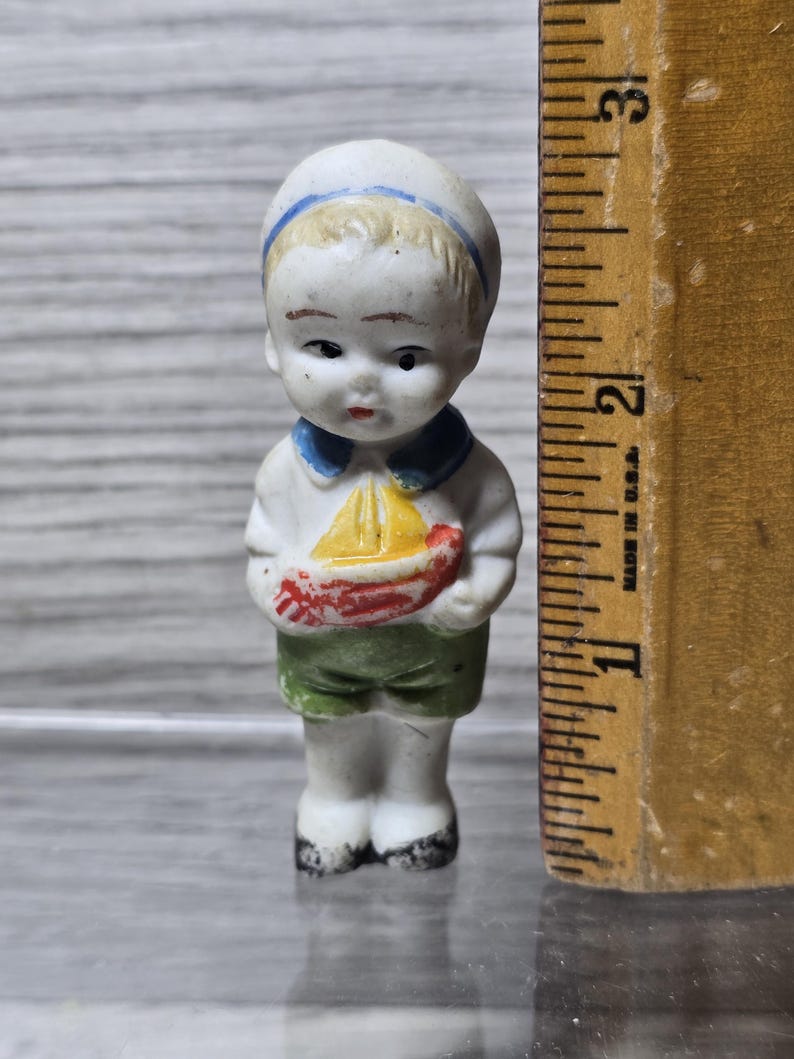 Pu&ograve; includere: Piccola statuina vintage in porcellana di un marinaio. La statuina indossa un cappello e una camicia bianchi con colletto blu, pantaloncini verdi e tiene in mano una barca rossa e gialla. La statuina &egrave; alta circa 6,35 cm.