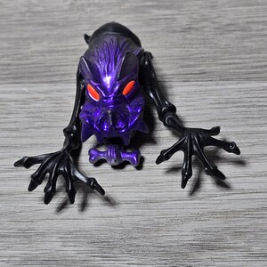 Op de afbeelding: Een paars en zwart monsterbeeldje met rode ogen. Het beeldje heeft een gedetailleerde kop, lange armen en klauwachtige handen. De figuur poseert alsof hij kruipt. Het speelgoed is gemaakt van plastic.