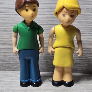 May include: Two plastic toy figures standing side by side. The figure on the left has brown hair, a green polo shirt, and blue pants. The figure on the right has yellow hair, a yellow sleeveless top, and a matching yellow skirt. Both have black shoes.
