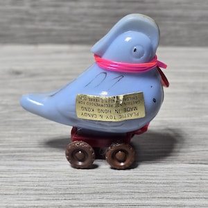 May include: A pale blue plastic bird toy, featuring a pink ribbon and a red base with brown wheels. The toy has a sticker that reads "PLASTIC TOY & CANDY MADE IN HONG KONG CAUTION NOT RECOMMENDED FOR CHILDREN UNDER 3 YEARS OLD."