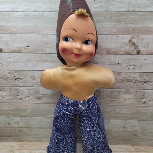 May include: A vintage doll with a brown and blue patterned hat, a gold-colored torso, and blue and white patterned pants. The doll has a smiling face with blue eyes and rosy cheeks.