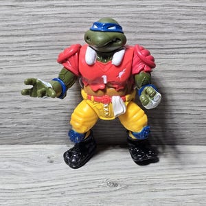 以下が含まれることがあります： アメフトのユニフォームを着たミュータント・タートルズのアクションフィギュア。フィギュアは緑色で青いマスクをしており、背番号1の赤いジャージを着ています。黄色のパンツ、黒い靴、青い手首と膝のパッドがルックを完成させています。
