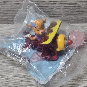 May include: A small, brown bear figurine in a purple shirt and blue bow riding in a red, yellow, and brown airplane toy. The airplane has a pink propeller and is packaged in a clear plastic bag.