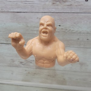 May include: A tan-colored plastic figure of a man with a muscular torso and arms. The figure is posed with its arms outstretched and its mouth open in a yell. The figure is made of a soft, pliable plastic.