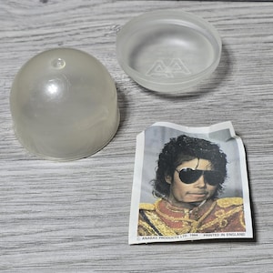 May include: A clear, plastic capsule with a matching lid, alongside a small paper image of Michael Jackson wearing sunglasses and a red and gold jacket. The image is printed in England.