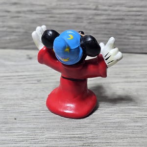 May include: A figurine of Mickey Mouse as the Sorcerer's Apprentice from Fantasia. The figure is seen from the back, wearing a red robe, a blue hat with yellow crescent moons and stars, and white gloves. The figure is posed with arms raised.