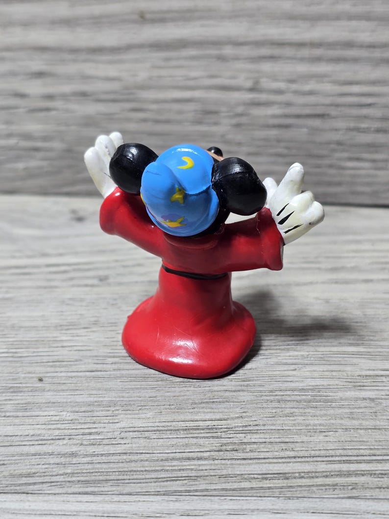 May include: A figurine of Mickey Mouse as the Sorcerer's Apprentice from Fantasia. The figure is seen from the back, wearing a red robe, a blue hat with yellow crescent moons and stars, and white gloves. The figure is posed with arms raised.