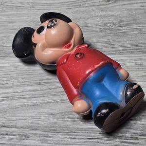 May include: A vintage Mickey Mouse figure, with a red jacket, blue trousers, and black shoes. The figure has a tan face, black ears, and a black hat. The toy is lying on a grey wooden surface.