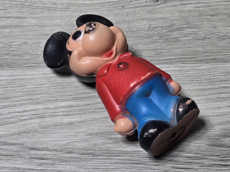 May include: A vintage Mickey Mouse figure, with a red jacket, blue trousers, and black shoes. The figure has a tan face, black ears, and a black hat. The toy is lying on a grey wooden surface.