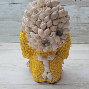 May include: A small, yellow and white seashell dog figurine. The dog is made of seashells and has a yellow yarn body.