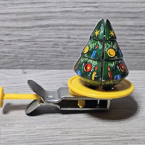 May include: A vintage Christmas tree toy with a green, decorated tree on a yellow base. The tree features colorful ornaments and candles. The toy has a silver and yellow mechanical base.