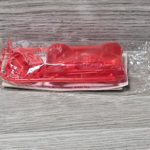 May include: A red plastic model car kit, still in its original clear plastic packaging. The kit includes various car parts and is set against a light gray wood-grain background.
