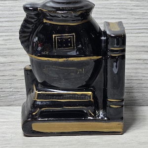 Puede incluir: Dos sujetalibros de cerámica negra con forma de estufa de leña con detalles dorados. La estufa tiene un cuerpo redondo con una chimenea y una base con una puerta. Los sujetalibros son un elemento decorativo para una biblioteca en casa.