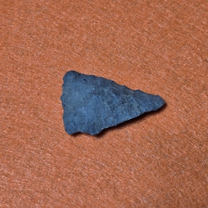 May include: A dark grey, triangular arrowhead with a rough texture. The stone artifact is set against an orange background, highlighting its shape and colour. The arrowhead is a historical artefact.