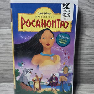 Könnte beinhalten: Eine Walt Disney Meisterwerk Pocahontas VHS-Kassette in einer weißen Kunststoffhülle. Das Cover zeigt Pocahontas, einen Waschbären, einen Kolibri und weitere Figuren. Der Text umfasst A Walt Disney Masterpiece und Pocahontas.