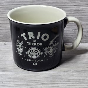 Puede incluir: Taza de cer&aacute;mica negra con interior y asa blancos. La taza presenta el texto "TRIO OF TERROR" e ilustraciones de personajes de "Oogie's Crew". La taza es de color oscuro.