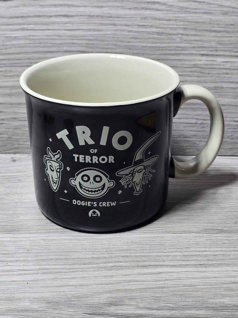 Puede incluir: Taza de cer&aacute;mica negra con interior y asa blancos. La taza presenta el texto "TRIO OF TERROR" e ilustraciones de personajes de "Oogie's Crew". La taza es de color oscuro.