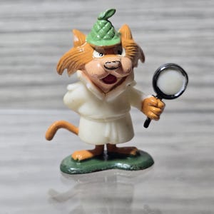 May include: A small orange cat figurine dressed as a detective, wearing a green deerstalker hat and a white trench coat. The cat holds a black-rimmed magnifying glass in one hand and stands on a green base, featuring a tail and pointed ears.