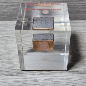 Puede incluir: Un bloque acrílico transparente con dos piezas metálicas rectangulares incrustadas. La pieza superior es plateada y la inferior es de color cobre. El texto "Wall-Tex Key Dealer" está impreso en rojo en la parte superior del bloque.