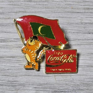 May include: A vintage pin featuring a cartoon tiger wearing a hat, holding a flag with a red background and a green crescent moon. The pin also includes a red Coca-Cola sign with gold lettering.