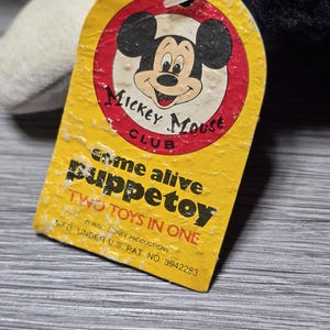 May include: Yellow tag with a red circle featuring Mickey Mouse's face. The tag reads "Mickey Mouse Club" and "Come alive puppetoy Two toys in one" with the text "WALT DISNEY PRODUCTIONS MFG. UNDER U.S. PAT. NO. 3942283".