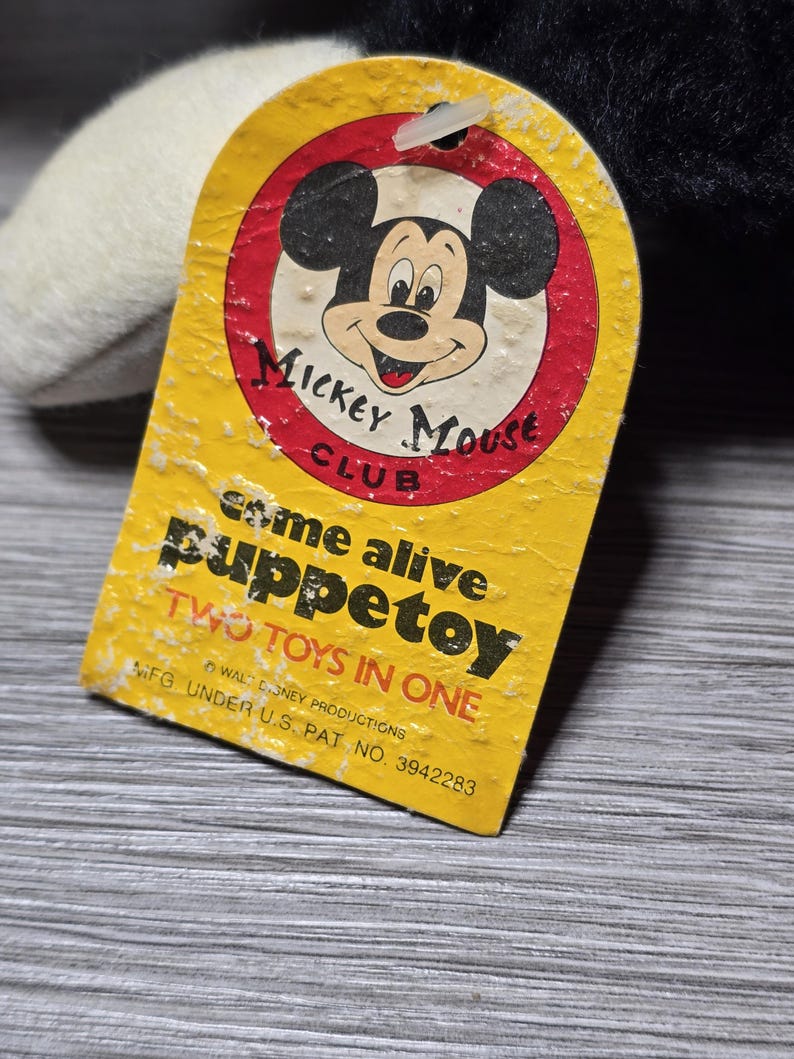 May include: Yellow tag with a red circle featuring Mickey Mouse's face. The tag reads "Mickey Mouse Club" and "Come alive puppetoy Two toys in one" with the text "WALT DISNEY PRODUCTIONS MFG. UNDER U.S. PAT. NO. 3942283".