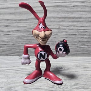 Può includere: Una statuetta di un personaggio dei cartoni animati rosso con lunghe antenne, un viso beige e guanti bianchi. Il personaggio ha un emblema circolare sul petto con una lettera N nera e tiene in mano una bomba sferica nera con una N bianca.