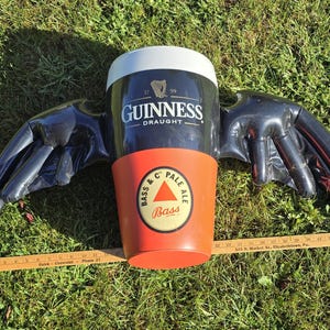 May include: A black and red inflatable Guinness beer mug with wings. The mug has the words "Guinness Draught" and a harp logo. The wings are black and have a textured surface. The mug is sitting on a green grassy surface.