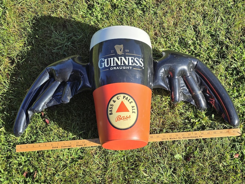 May include: A black and red inflatable Guinness beer mug with wings. The mug has the words "Guinness Draught" and a harp logo. The wings are black and have a textured surface. The mug is sitting on a green grassy surface.
