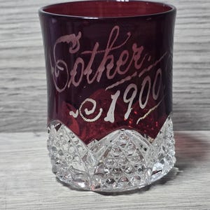 Könnte beinhalten: Ein zylindrisches Glas mit einem tiefroten, cranberryfarbenen oberen Bereich, der die geätzte weiße Schrift "Sother" und "1900" aufweist. Der untere Teil des Glases ist klar und mit einem strukturierten Rautenmuster am Boden versehen.