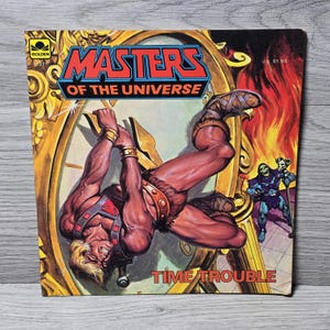 Puede incluir: Una portada de c&oacute;mic vintage de "Masters of the Universe". La portada presenta un personaje musculoso colgando de un reloj, con el t&iacute;tulo "Time Trouble". El fondo incluye una esfera de reloj, llamas y otros personajes.