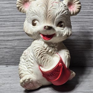 Peut inclure: Figurine d'ours blanc vintage avec un pot de miel rouge. L'ours a un visage souriant, des yeux bruns et des accents roses sur les oreilles. Le pot de miel rouge porte l'inscription "HONEY". La figurine est posée sur une surface en bois grise.