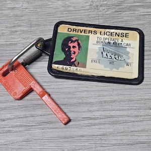 May include: A novelty keychain featuring a rectangular plastic tag designed to look like a driver's license for a Burnin' Key Car, paired with a bright orange plastic key-shaped fob attached by a metal carabiner clip on a black plastic frame.