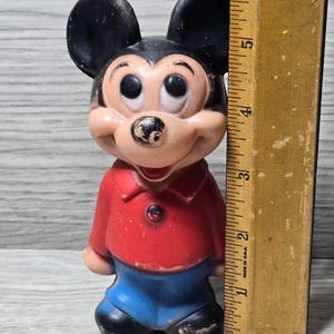 May include: Vintage Mickey Mouse figurine. The figure has black ears, a tan face, and is wearing a red shirt and blue shorts. A wooden ruler is next to the figurine, measuring approximately 15 cm.