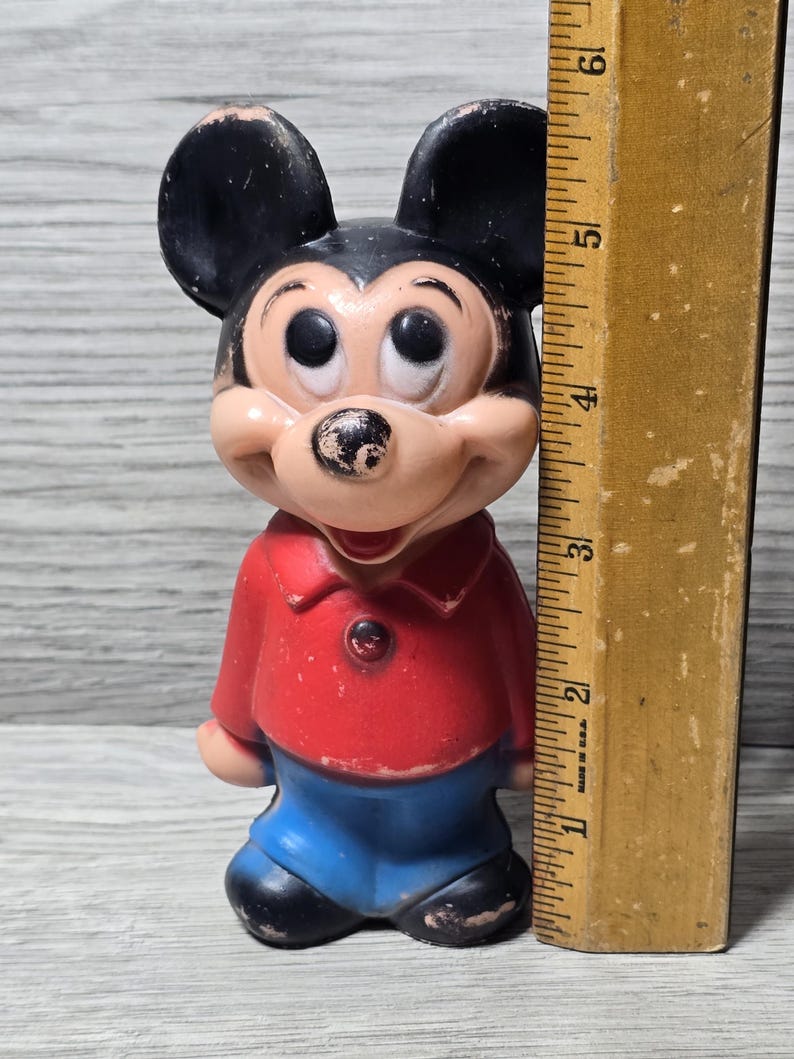 May include: Vintage Mickey Mouse figurine. The figure has black ears, a tan face, and is wearing a red shirt and blue shorts. A wooden ruler is next to the figurine, measuring approximately 15 cm.