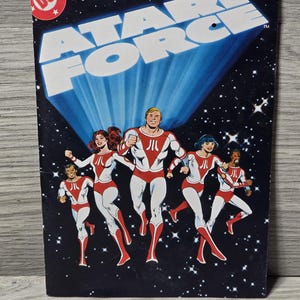 Può includere: Una copertina di fumetto vintage con il titolo "ATARI FORCE" in lettere bianche su uno sfondo spaziale blu e nero. La copertina mostra un gruppo di cinque figure in costume rosso e bianco, con il logo DC Comics.