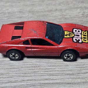 May include: A red die-cast toy sports car featuring a black interior, black wheels, and a yellow Ferrari logo on the hood. The hood displays the text 308 G.T.B. in bold black and white lettering. The car shows signs of wear with minor paint chips.