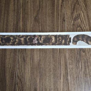 Puede incluir: Una pegatina rectangular con un patrón de pelaje marrón y negro y un diseño de cola separada. La pegatina está sobre un fondo blanco.