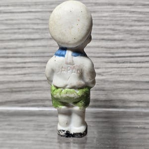 Pu&ograve; includere: Piccola statuina vintage in ceramica di un ragazzo in tenuta da marinaio. La statuina &egrave; bianca con colletto blu, pantaloncini verdi e la scritta "JAPAN" sul retro. La statuina &egrave; in piedi con le braccia incrociate.
