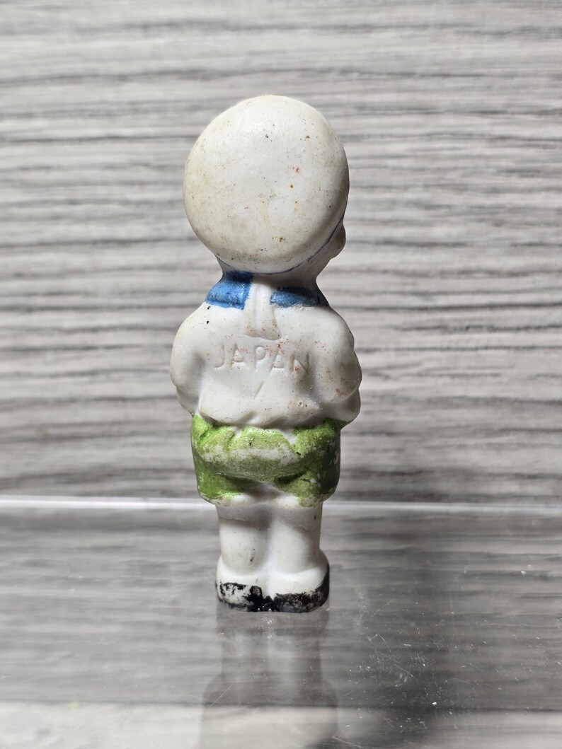 Pu&ograve; includere: Piccola statuina vintage in ceramica di un ragazzo in tenuta da marinaio. La statuina &egrave; bianca con colletto blu, pantaloncini verdi e la scritta "JAPAN" sul retro. La statuina &egrave; in piedi con le braccia incrociate.