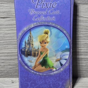 Pode incluir: Uma caixa roxa para uma coleção de moedas prensadas Pixie, com uma imagem de Tinkerbell em um vestido verde e um castelo. A caixa tem uma borda prateada brilhante e as palavras "Pixie Pressed Coin Collection".