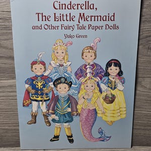May include: A light blue book cover featuring the title "Cinderella, The Little Mermaid and Other Fairy Tale Paper Dolls". The cover displays illustrations of paper dolls in various fairy tale costumes, including a mermaid and a princess.