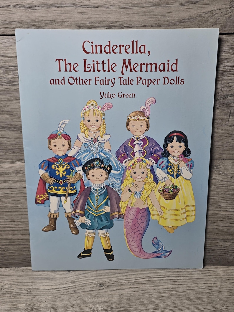 May include: A light blue book cover featuring the title "Cinderella, The Little Mermaid and Other Fairy Tale Paper Dolls". The cover displays illustrations of paper dolls in various fairy tale costumes, including a mermaid and a princess.
