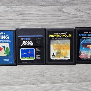 May include: Four vintage Atari video game cartridges. Titles include "Skiing," "Space Jockey," "Haunted House," and "Space Invaders." The cartridges are black with colorful labels featuring game graphics and text. The games are displayed on a wooden surface.