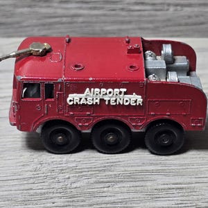 May include: A vintage red die-cast metal toy fire truck with the words "AIRPORT CRASH TENDER" in white lettering. The toy has six black wheels and a gold-colored nozzle on top.